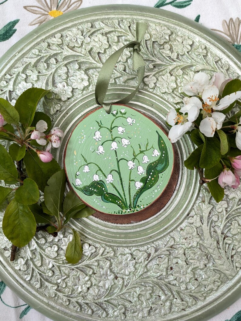 May include: A round, wooden ornament with a hand-painted lily of the valley design. The ornament is painted on a light green background and has a brown border. A green ribbon is attached for hanging. The ornament is surrounded by fresh flowers and leaves.