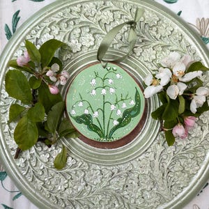 May include: A round, hand-painted wooden ornament featuring a lily of the valley design on a green background. The ornament is accented with a green ribbon and rests on a decorative, light green, floral-patterned plate. Fresh flowers and greenery surround the ornament.