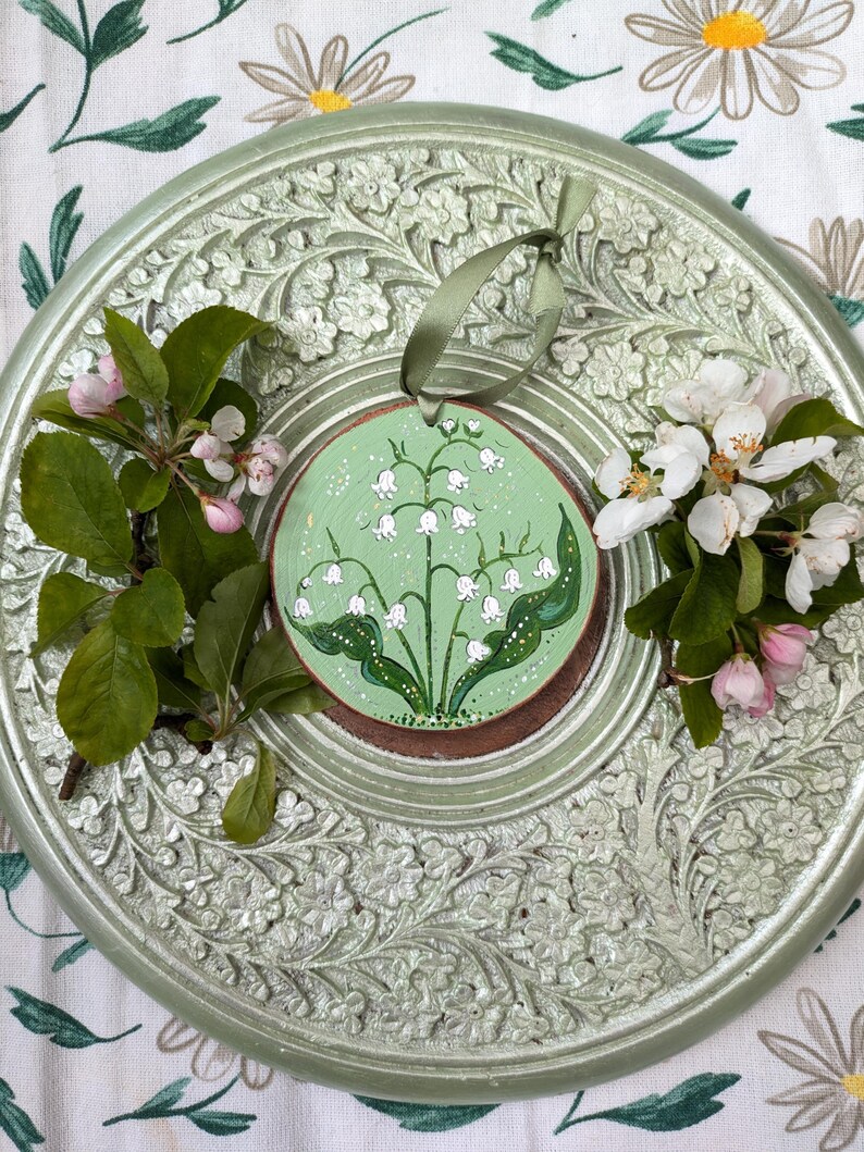 May include: A round, hand-painted wooden ornament featuring a lily of the valley design on a green background. The ornament is accented with a green ribbon and rests on a decorative, light green, floral-patterned plate. Fresh flowers and greenery surround the ornament.