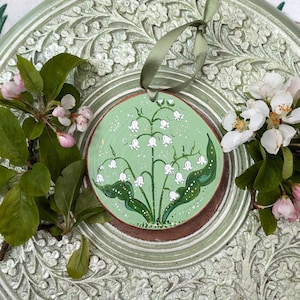 May include: A round, wooden ornament featuring a hand-painted lily of the valley design on a light green background. The ornament has a brown border and a green ribbon for hanging. The ornament is surrounded by green leaves and white flowers.
