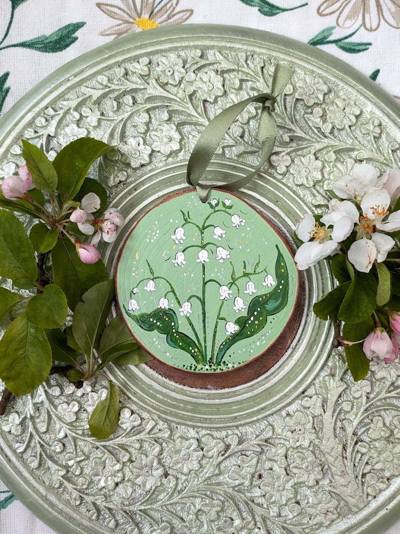 May include: A round, wooden ornament featuring a hand-painted lily of the valley design on a light green background. The ornament has a brown border and a green ribbon for hanging. The ornament is surrounded by green leaves and white flowers.