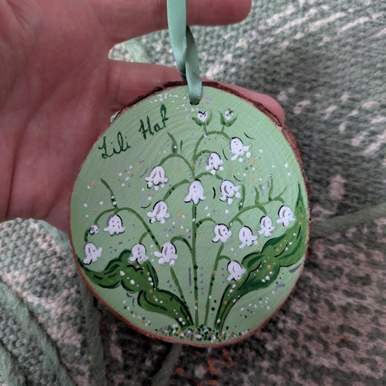 May include: Hand-painted wooden ornament featuring a lily of the valley design. The round ornament has a light green background with white flowers and green leaves. The text "Lili Haf" is written in cursive. A light green ribbon is attached for hanging.