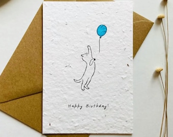 May include: A white textured paper greeting card featuring a black line drawing of a cat reaching for a blue balloon. The text Happy Birthday is printed in black ink at the bottom. The card is placed on a brown paper envelope.