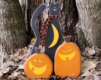 May include: Black cat with a red and black plaid scarf and a yellow crescent moon on its chest, standing between two orange pumpkins with yellow faces. The pumpkins and cat are wooden and painted. The scene is set in a forest with brown leaves on the ground.