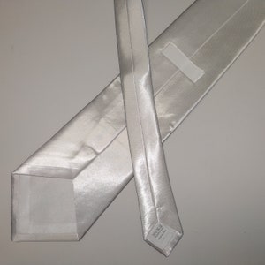 May include: A light silver-colored necktie, made of a shiny, smooth fabric. The tie is laid out flat, showing the front and back, with a square end and a pointed end. The fabric has a subtle sheen, reflecting light.