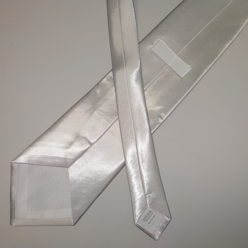 May include: A light silver-colored necktie, made of a shiny, smooth fabric. The tie is laid out flat, showing the front and back, with a square end and a pointed end. The fabric has a subtle sheen, reflecting light.