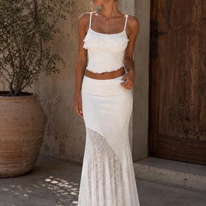 Puede incluir: Un conjunto de dos piezas blanco que consta de un top corto tipo camisola con escote de volantes y una falda larga a juego. La falda presenta un corte de cintura alta y un panel asimétrico de encaje floral detallado en la parte inferior.