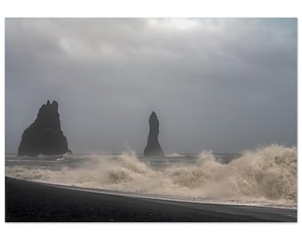 May include: A landscape photograph of a black sand beach with large, dark, jagged rock formations rising from the ocean. White sea foam crashes against the shore under a heavy, overcast, grey sky, creating a moody and atmospheric coastal scene.