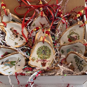 May include: A set of twelve oyster shell ornaments arranged in a cardboard box with shredded paper filler. Each shell features a unique Christmas-themed illustration, gold-painted edges, and a red ribbon hanger with gold and red beads for display.