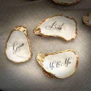 May include: Several oyster shells with gold-painted edges are arranged on a white surface. Each shell has a handwritten word in black script, including "Bride," "Groom," "Mr. & Mrs.," "Bridesmaid," and "Groom's Mom." The shells are wedding-themed.