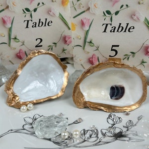 May include: Two white oyster shells with gold trim, used as table number holders. The shells are sitting on a white plate with a floral design. The table numbers are on cards with floral designs and the text "Table 2" and "Table 5".
