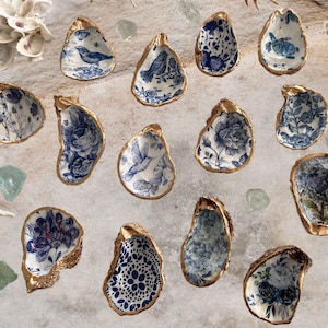 May include: A collection of oyster shell dishes with gold rims and blue and white floral or bird designs. The shells are arranged on a light-colored surface with seashells and sea glass accents, creating a coastal theme.