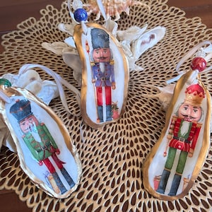 May include: Three oyster shell Christmas ornaments featuring nutcracker soldier designs. Each ornament has a gold-painted edge, a ribbon for hanging, and a decorative bead. The nutcrackers are dressed in red, green, and purple uniforms.