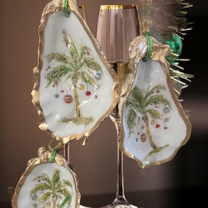 May include: Hand-painted oyster shell ornaments with a gold-painted edge. Each shell features a palm tree decorated with colorful ornaments, and a green ribbon for hanging. A festive, tropical Christmas decoration.