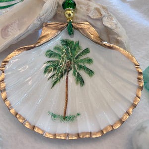 May include: A handcrafted seashell ornament featuring a painted palm tree. The white shell has a gold-painted edge and is adorned with a green and gold beaded top. The palm tree is painted in green and brown.