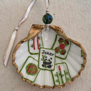 May include: A hand-painted seashell ornament with a gold-painted edge. The ornament features a Mahjong tile design with a panda and the word "Joker". A green bead and white ribbon complete the design. The text "#4 Green" is at the bottom.