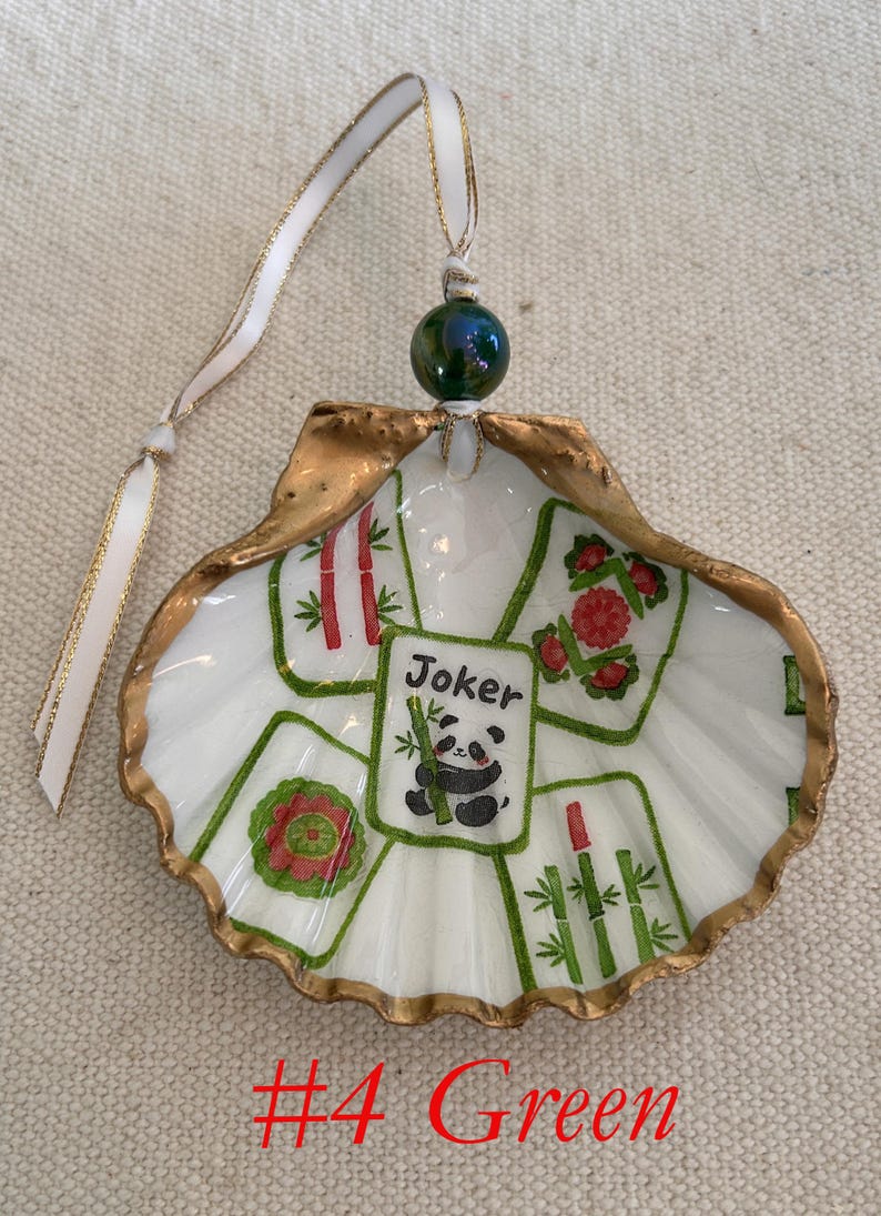 May include: A hand-painted seashell ornament with a gold-painted edge. The ornament features a Mahjong tile design with a panda and the word "Joker". A green bead and white ribbon complete the design. The text "#4 Green" is at the bottom.