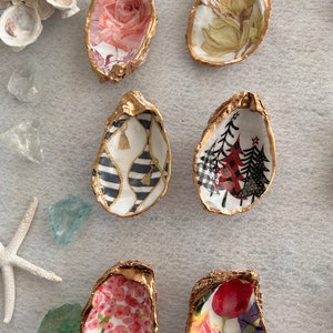 May include: Six gold oyster shell dishes with different floral and patterned designs. The designs include a pink rose, a green floral pattern, a black and white striped pattern with tassels, a red and black Christmas tree pattern, a pink floral pattern, and a red and yellow floral pattern.