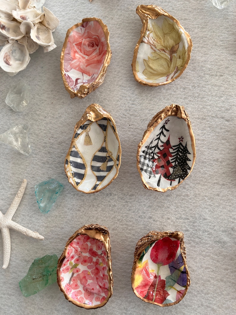 May include: Six gold oyster shell dishes with different floral and patterned designs. The designs include a pink rose, a green floral pattern, a black and white striped pattern with tassels, a red and black Christmas tree pattern, a pink floral pattern, and a red and yellow floral pattern.