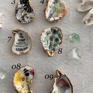 May include: Nine oyster shells with gold trim, each painted with a different design. The designs include a tropical leaf, a bird, a Christmas tree, a floral design, a bee, and a flamingo. The shells are numbered 3, 4, 7, 8, 08, and 09.