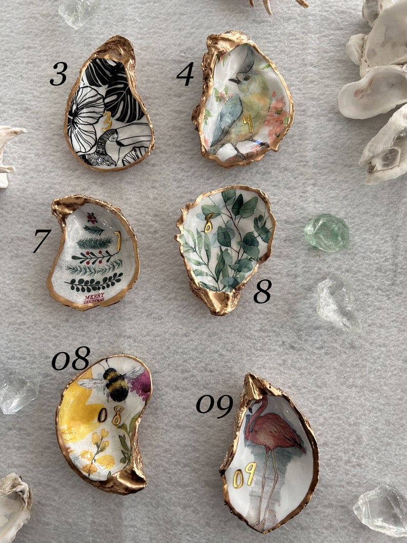 May include: Nine oyster shells with gold trim, each painted with a different design. The designs include a tropical leaf, a bird, a Christmas tree, a floral design, a bee, and a flamingo. The shells are numbered 3, 4, 7, 8, 08, and 09.