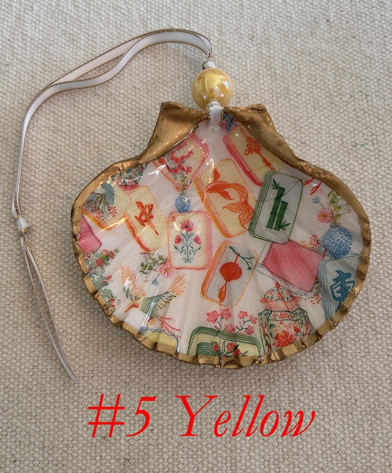 May include: A decorative seashell ornament with a gold-painted edge, featuring a colorful design of floral and card motifs. It has a white ribbon for hanging and a yellow bead at the top. The text "#5 Yellow" is visible.