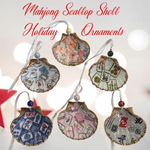 May include: Mahjong Scallop Shell Holiday Ornaments. Six seashell ornaments with gold trim, each featuring a unique Mahjong tile design. The shells are suspended by white ribbons and adorned with decorative beads. The text "Mahjong Scallop Shell Holiday Ornaments" is displayed in red.