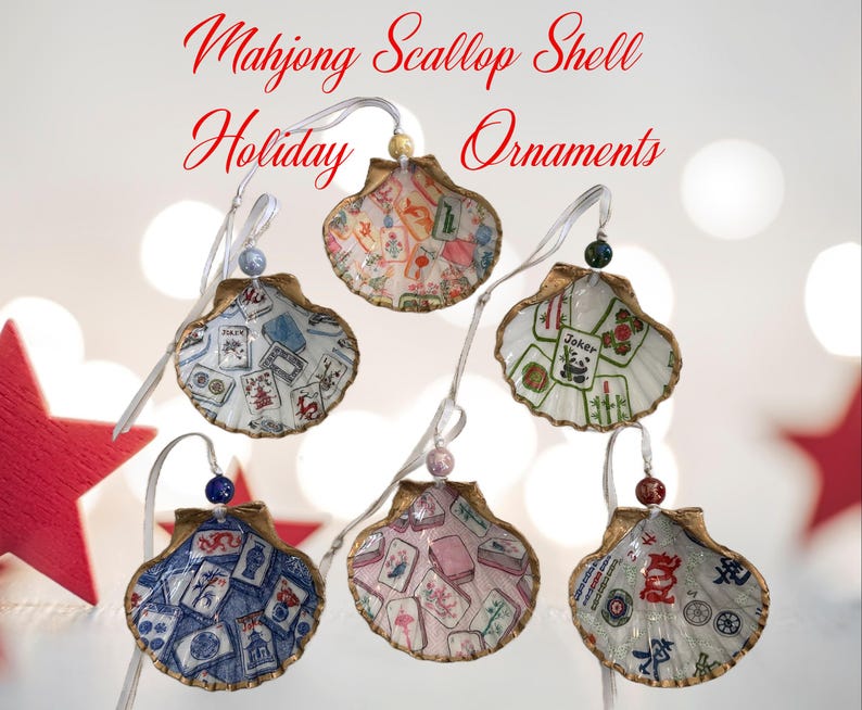May include: Mahjong Scallop Shell Holiday Ornaments. Six seashell ornaments with gold trim, each featuring a unique Mahjong tile design. The shells are suspended by white ribbons and adorned with decorative beads. The text "Mahjong Scallop Shell Holiday Ornaments" is displayed in red.