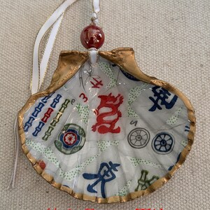 May include: A seashell ornament with a gold-painted edge, featuring a printed game tile design in red, blue, and green. The ornament has a white ribbon with gold trim and a red bead for hanging. The text "#6 Game Tile" is visible.