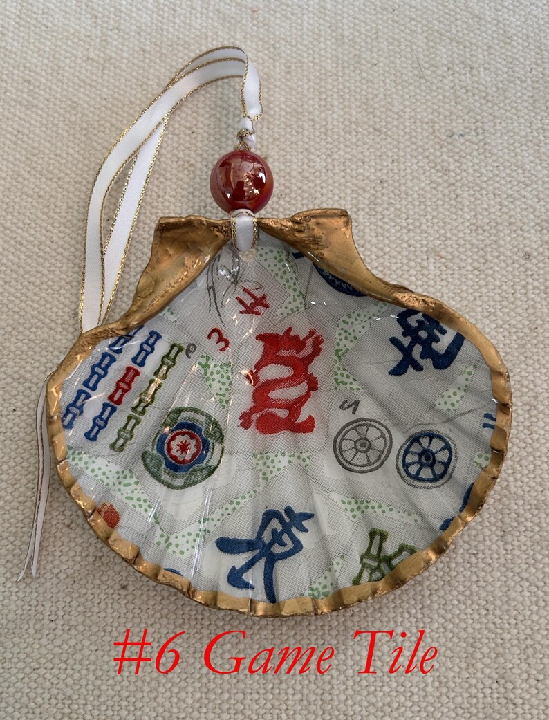 May include: A seashell ornament with a gold-painted edge, featuring a printed game tile design in red, blue, and green. The ornament has a white ribbon with gold trim and a red bead for hanging. The text "#6 Game Tile" is visible.