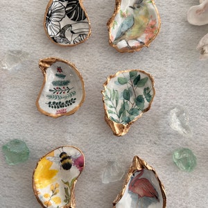 May include: Six oyster shells with gold trim, each decorated with a different design. The designs include a toucan, a bird, a Christmas tree, eucalyptus leaves, a bee and flowers, and a flamingo.