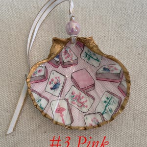 May include: A decorative seashell ornament with a gold-painted edge, featuring a pink interior with printed images of Mahjong tiles. The ornament is adorned with a white ribbon with a gold edge and a pink bead. The text "#3 Pink" is visible.