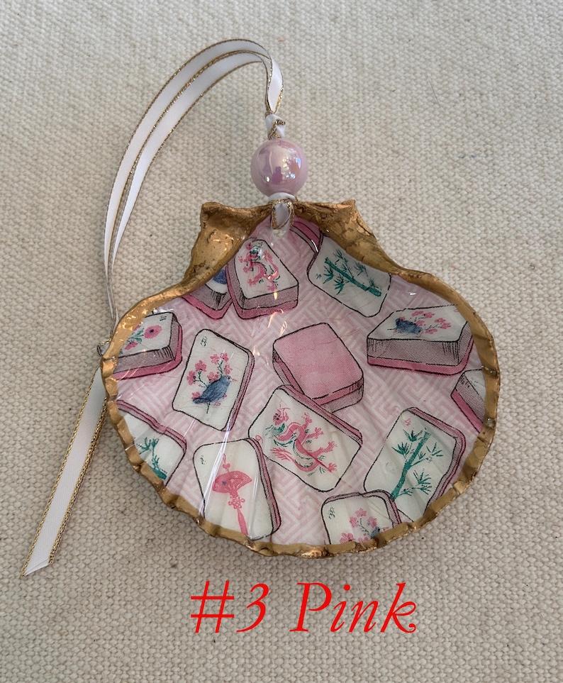 May include: A decorative seashell ornament with a gold-painted edge, featuring a pink interior with printed images of Mahjong tiles. The ornament is adorned with a white ribbon with a gold edge and a pink bead. The text "#3 Pink" is visible.