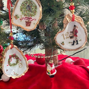 May include: Three oyster shell ornaments hanging from a tree. One features a partridge in a pear tree with the number 1, another shows a drummer with the number 12, and the third displays a wreath with the number 5. Each has gold-painted edges.