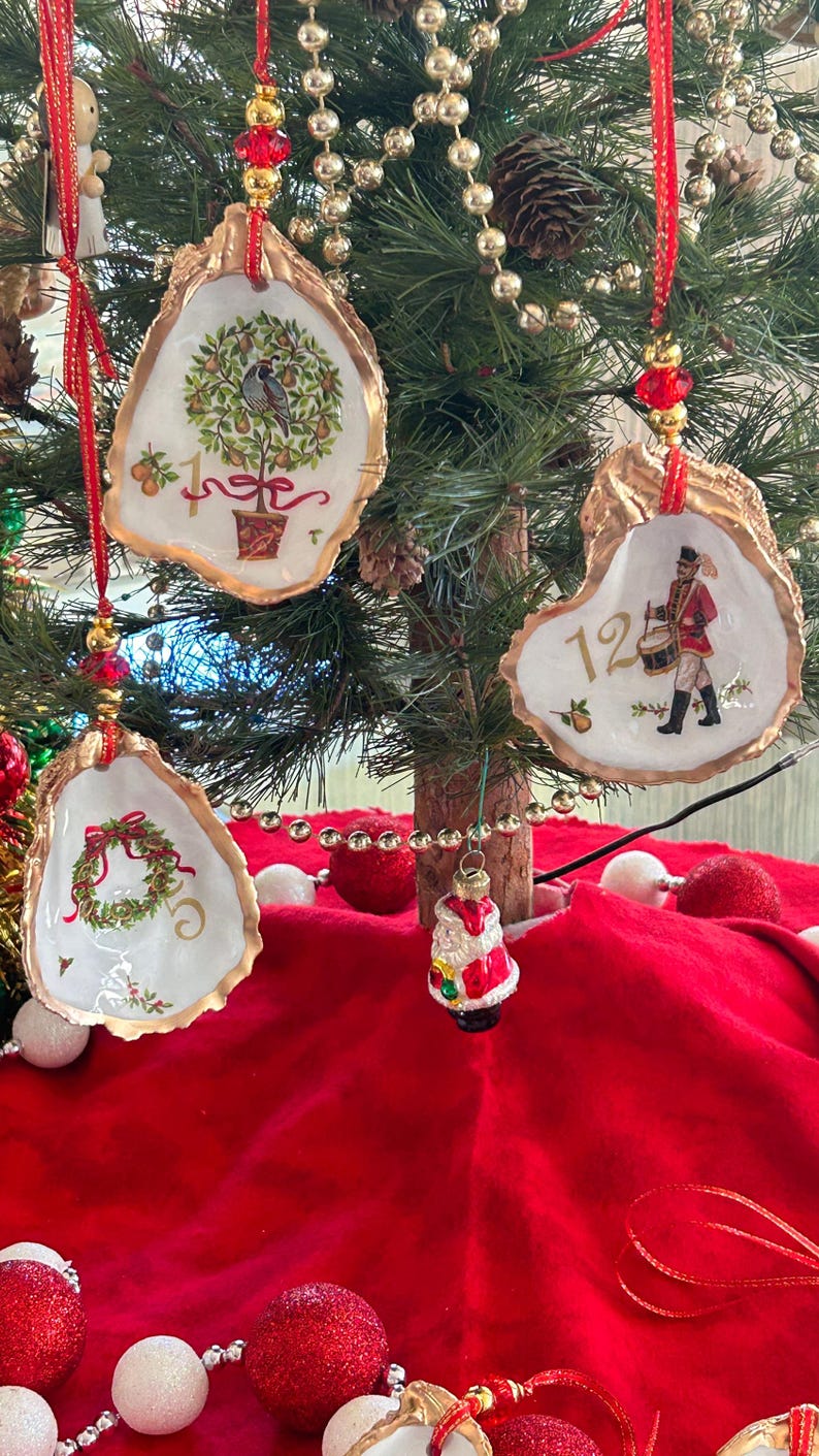 May include: Three oyster shell ornaments hanging from a tree. One features a partridge in a pear tree with the number 1, another shows a drummer with the number 12, and the third displays a wreath with the number 5. Each has gold-painted edges.