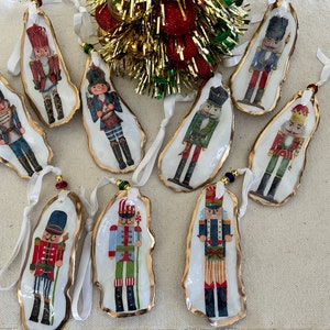 May include: A collection of ten oyster shell Christmas ornaments, each featuring a different nutcracker soldier design. The shells have gold-painted edges and are strung with white ribbon and decorative beads. The ornaments are a festive holiday decoration.