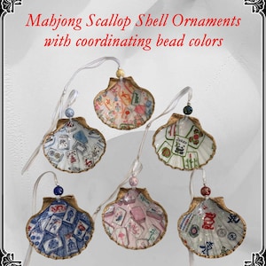 May include: Six scallop shell ornaments with coordinating bead colors. Each shell is painted with a different Mahjong tile design. The shells are trimmed in gold and have a white ribbon for hanging. The text "Mahjong Scallop Shell Ornaments with coordinating bead colors" is at the top.