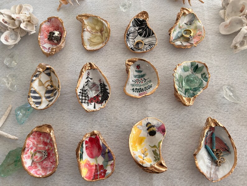May include: Twelve oyster shell ring dishes with gold trim. The dishes are decorated with various designs including floral patterns, Christmas trees, and a bee. Some of the dishes have a ring inside them.