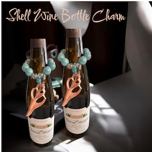 May include: Two wine bottles adorned with shell-shaped charms. Each bottle features a beaded necklace in shades of turquoise and clear beads, with a shell pendant. The text "Shell Wine Bottle Charm" is at the top.