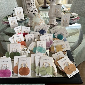 May include: Assortment of colorful shell earrings displayed on cards with the text "Tides of Time" on each card. The earrings are made of natural seashells and have gold-toned hoops.