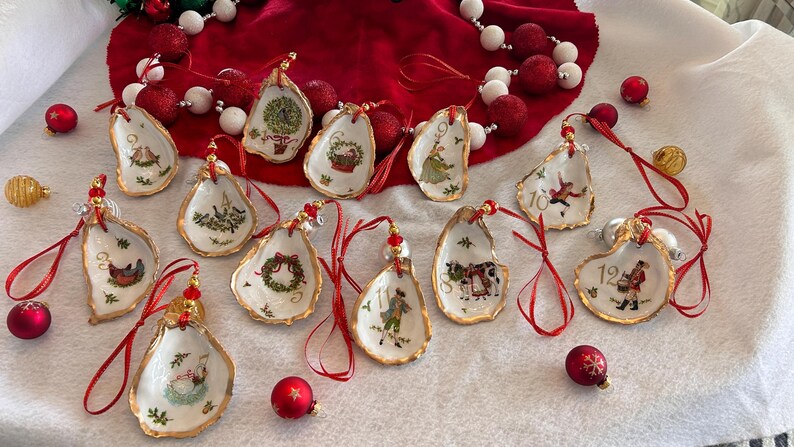 May include: A set of twelve oyster shell ornaments, each featuring a unique hand-painted Christmas illustration and a gold-painted edge. Each shell is numbered from one to twelve and includes a red ribbon loop for hanging, accented with small red beads.
