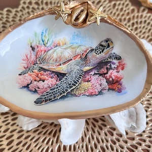 May include: A decorative seashell dish with a painted sea turtle design. The turtle rests on a bed of coral in shades of pink, red, and purple. The shell has a gold-painted rim and is adorned with starfish and a small shell.