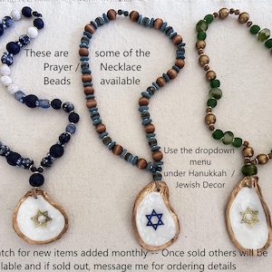May include: Three beaded necklaces with oyster shell pendants. The necklaces feature blue, brown, and green beads. Each pendant has a Star of David design. Text on the image says "Prayer Beads" and "Necklace available."