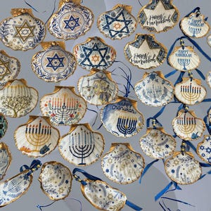 May include: A collection of hand-painted seashell ornaments with gold trim, featuring Jewish symbols like the Star of David and menorahs. Some shells have "Happy Hanukkah" text. Blue ribbons are attached for hanging.
