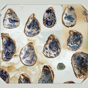 May include: A collection of decorative oyster shells with gold-painted edges. Each shell features a unique blue and white porcelain design, including birds, flowers, and other patterns. The shells are arranged against a soft, blurred background.