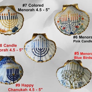 May include: A set of three ceramic mugs with a speckled, cream-colored glaze. Each mug features a different hand-painted design: a blue and green leaf, a yellow flower, and a red berry. The mugs have a simple, modern design.
