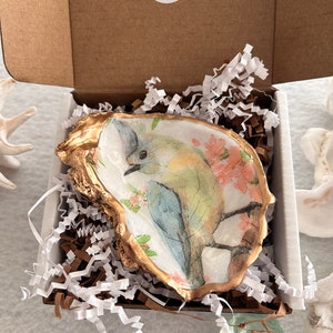 May include: A small, white oyster shell dish with a gold rim. The dish is decorated with a watercolor painting of a bluebird perched on a branch with pink flowers. The dish is in a white box with white shredded paper. A sticker on the box says 'Thank you' with two red hearts.