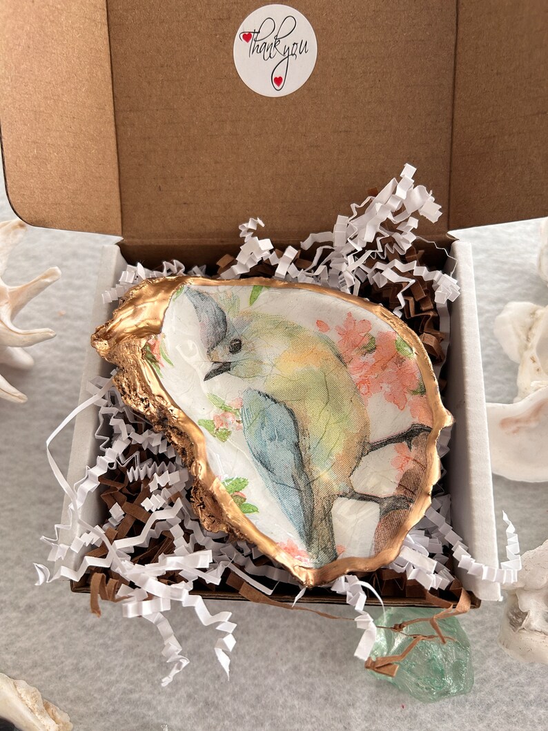 May include: A small, white oyster shell dish with a gold rim. The dish is decorated with a watercolor painting of a bluebird perched on a branch with pink flowers. The dish is in a white box with white shredded paper. A sticker on the box says 'Thank you' with two red hearts.