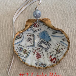 May include: A hand-painted seashell ornament with a gold-painted edge, featuring a light blue interior with illustrations of playing cards. The ornament has a white ribbon with gold trim and a light blue bead for hanging. The text "#2 Light Blue" is at the bottom.