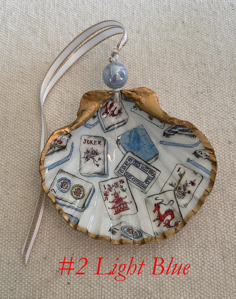 May include: A hand-painted seashell ornament with a gold-painted edge, featuring a light blue interior with illustrations of playing cards. The ornament has a white ribbon with gold trim and a light blue bead for hanging. The text "#2 Light Blue" is at the bottom.
